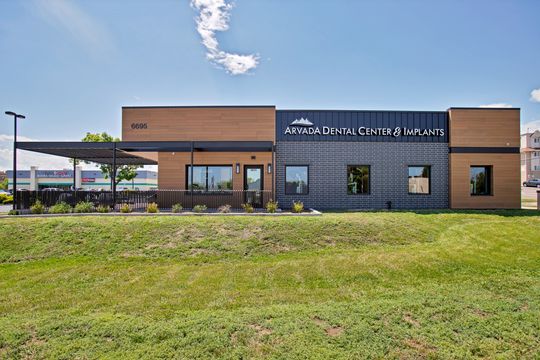 A full exterior street view of Arvada Dental Center & Implants in Colorado, highlighting the modern building design, large exterior signage, address number, patio area, and landscaped frontage.
