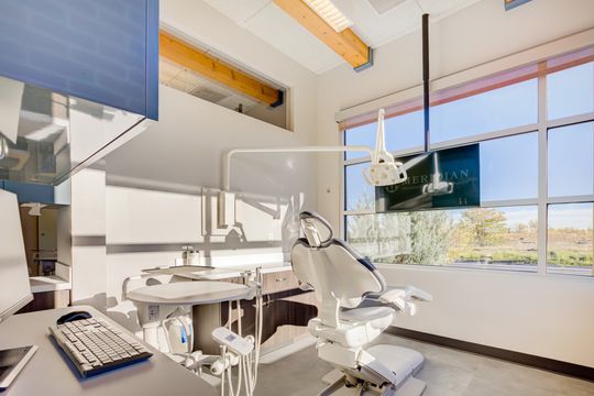 This image of the bright periodontal operatory at Meridian Periodontics of Central Park shows dental equipment, chair, large windows, wall monitor, and warm wood ceiling beams.