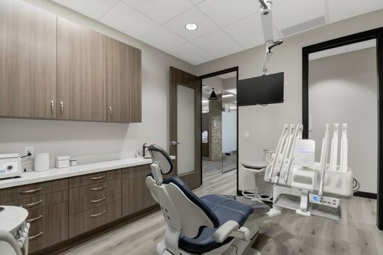 This image of the dental operatory with cabinetry at Soult and Swanson Family Dentistry shows a dental chair, wall monitor, brown cabinetry, sink station, and doorway to the clinical hall.