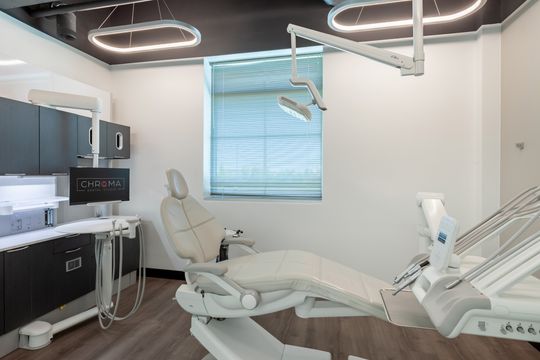 This image of the dental treatment room at Chroma Dental Studio shows a dental chair, overhead light, wall monitor, window shade, and integrated clinical cabinetry.