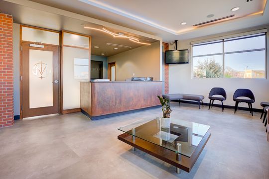 This image of the reception desk and waiting area at Meridian Periodontics of Central Park shows a stone reception counter, purple seating, coffee table, wall monitor, and window light.