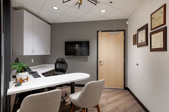 This image of the consultation room at Chroma Dental Studio shows a white meeting table, upholstered chairs, wall monitor, framed artwork, and warm wood flooring.