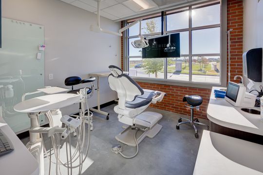 This image of the full treatment room at Meridian Periodontics of Central Park shows a dental chair, wall monitor, brick wall, large windows, and integrated clinical cabinetry.