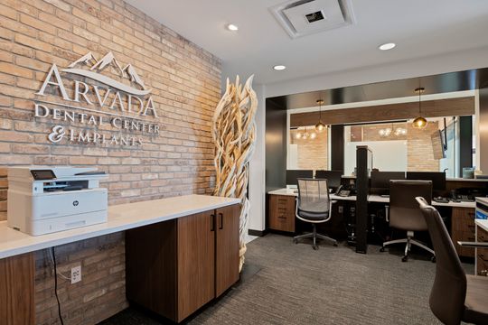 A front office view at Arvada Dental Center showing the branded brick wall, administrative workspace, wood cabinetry, printer station, and warm modern design details.