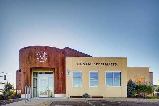 This image of the exterior front elevation at Meridian Periodontics of Central Park shows a modern dental specialists building, curved wood entry facade, signage, landscaping, and blue sky.