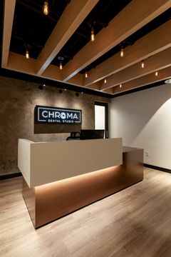 This image of the reception desk detail at Chroma Dental Studio shows a compact reception counter, Chroma logo wall, wood ceiling slats, and soft modern lighting.