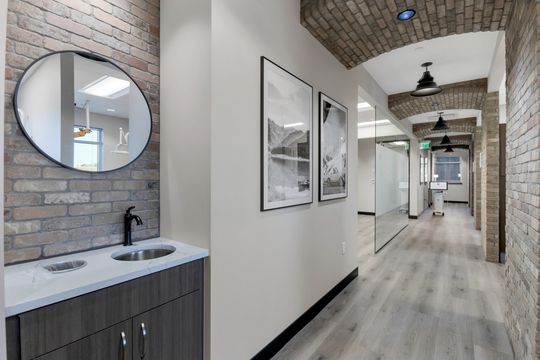 This image of the hallway sink and mirror detail at Soult and Swanson Family Dentistry shows a round mirror, sink cabinet, brick wall, framed art, and arched clinical corridor.