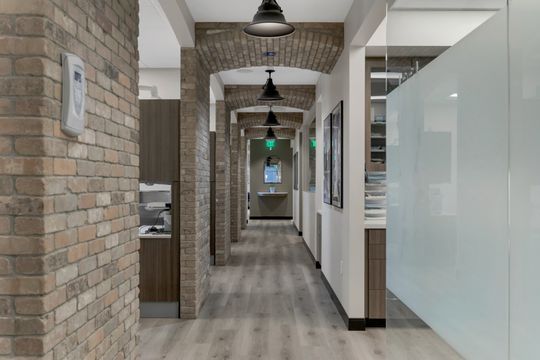 This image of the brick arch clinical hallway at Soult and Swanson Family Dentistry shows brick columns, glass partitions, operatory entrances, pendant lights, and wood-look flooring.