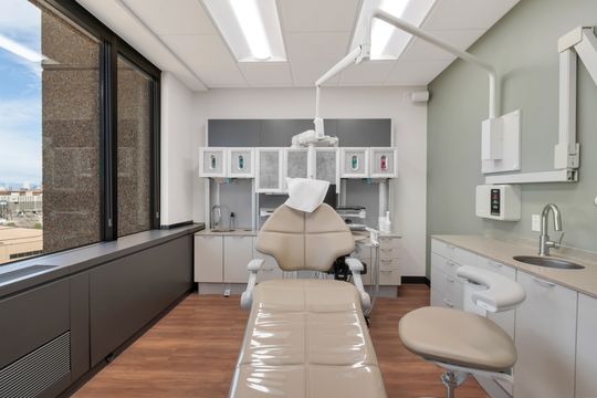 A clean patient treatment room at Bridge Creek Dentistry with a dental chair, overhead equipment, window light, clinical cabinetry, sink, and soft green wall accents.