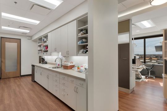 A bright dental lab workspace at Bridge Creek Dentistry featuring white cabinetry, open shelving, sink area, clinical supplies, and direct access to nearby treatment rooms.