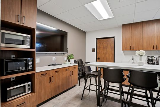 A modern staff break room at Arvada Dental Center featuring wood cabinetry, high-top seating, a large wall-mounted screen, kitchen appliances, coffee station, and clean contemporary finishes.