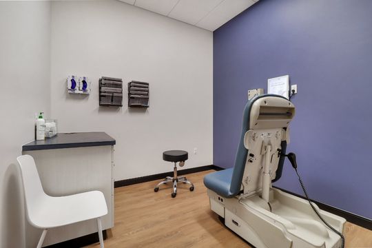 A clean dermatology exam room at Elevation Dermatology with a blue accent wall, exam chair, workstation, and patient seating.