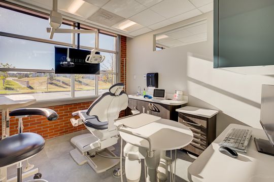 This image of the modern periodontal operatory at Meridian Periodontics of Central Park shows dental chair equipment, cabinetry, wall monitor, window light, and warm brick accents.