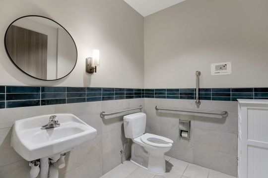 This image of the accessible restroom sink view at Soult and Swanson Family Dentistry shows round mirror, white sink, grab bars, blue tile accent, and neutral restroom walls.