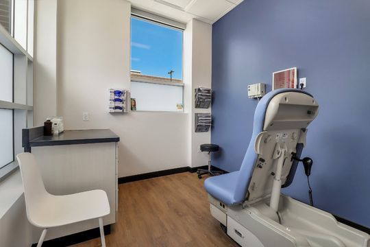 A dermatology exam room at Elevation Dermatology with natural light, blue accent wall, exam chair, counter space, and patient seating.