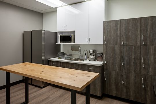 This image of the staff breakroom and storage area at Chroma Dental Studio shows a wood table, refrigerator, microwave, cabinetry, sink, and clean staff workspace.