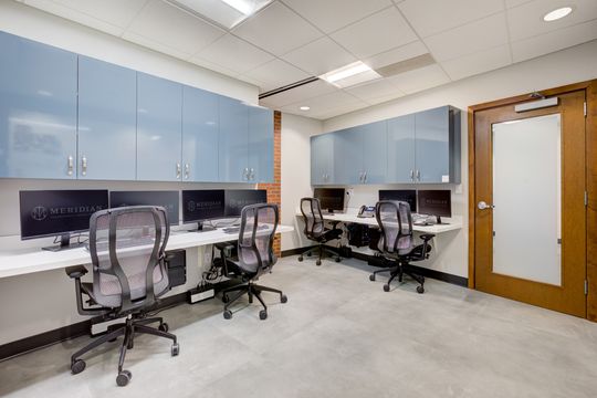 This image of the conference and team workroom at Meridian Periodontics of Central Park shows conference tables, task chairs, wall monitor, cabinetry, and bright office lighting.
