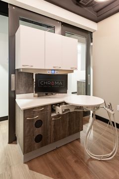 This image of the dental operatory detail at Chroma Dental Studio shows a dental chair, round work counter, cabinetry, sink, wall monitor, and treatment tools.