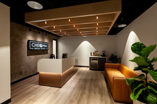 This image of the reception desk and lounge at Chroma Dental Studio shows a wood and white reception desk, illuminated logo wall, ceiling slats, orange seating, and indoor greenery.