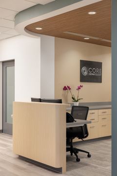 This image of the reception workstation detail at Core Dermatology shows a compact front desk, Core Dermatology wall sign, floral accent, and curved wood ceiling detail.