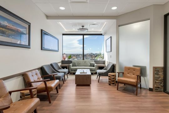 A comfortable patient lounge at Bridge Creek Dentistry featuring a green sofa, leather chairs, wood flooring, artwork, large windows, and a calm waiting environment.