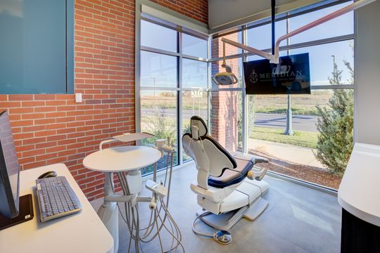 This image of the periodontal treatment room at Meridian Periodontics of Central Park shows a dental chair, wall monitor, large windows, exposed brick, and clinical cabinetry.