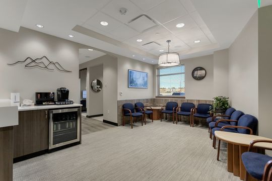 This image of the patient lounge and coffee bar at Soult and Swanson Family Dentistry shows blue waiting chairs, coffee station, pendant lighting, window seating, and neutral reception finishes.