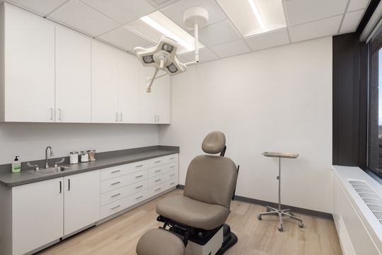 This image of the dermatology exam room at Core Dermatology shows an exam chair, overhead procedure light, white cabinetry, sink, and clean clinical counter space.