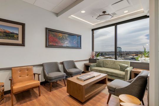 A relaxing waiting area at Bridge Creek Dentistry with lounge seating, large windows, mountain views, framed artwork, wood flooring, and warm patient-friendly design.
