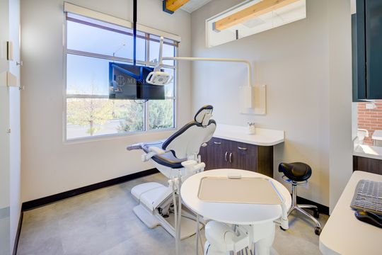 This image of the treatment room with dental chair at Meridian Periodontics of Central Park shows a dental chair, wall monitor, window light, white cabinetry, and round clinical work surface.