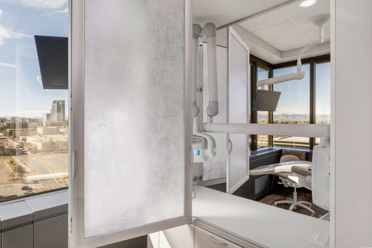 A window-side detail of a Bridge Creek Dentistry treatment room showing privacy glass, dental equipment, patient monitor, and an elevated city view.