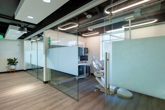This image of the glass hallway and treatment rooms at Chroma Dental Studio shows frosted glass walls, wood flooring, exposed ceiling, treatment room access, and modern lighting.