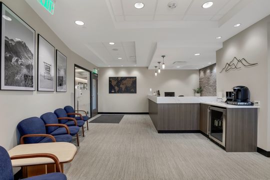 This image of the waiting room with reception desk at Soult and Swanson Family Dentistry shows blue chairs, wall art, front desk, coffee station, and bright lobby lighting.