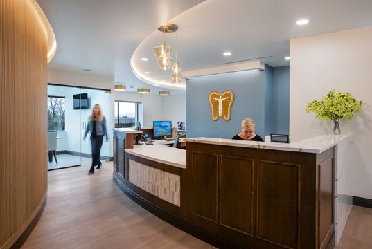 This image of the reception desk and logo wall at Collective Health shows front desk cabinetry, computer stations, illuminated tooth logo, curved ceiling light, and greenery.