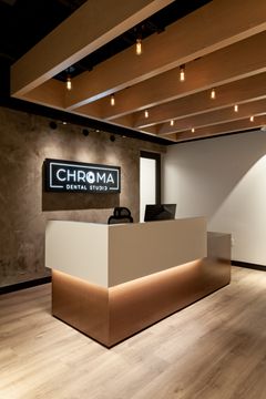 This image of the reception desk vertical view at Chroma Dental Studio shows a front desk, illuminated Chroma logo sign, warm ceiling slats, and modern neutral finishes.