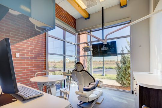 This image of the treatment room with brick wall at Meridian Periodontics of Central Park shows a dental chair, wall monitor, brick accent wall, round work surface, and landscape views.