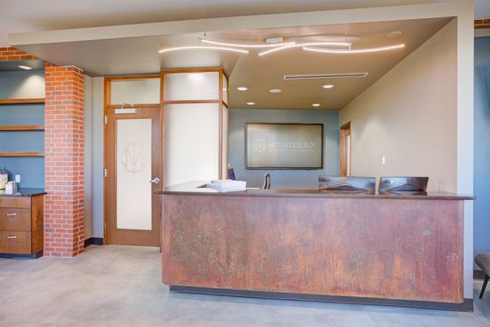 This image of the front desk counter at Meridian Periodontics of Central Park shows a stone reception counter, wood-framed windows, waiting chairs, and warm ceiling lighting.