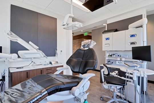 A contemporary dental operatory at Arvada Dental Center featuring a black patient chair, overhead dental light, treatment tools, computer workstation, wood cabinetry, and clean clinical finishes.