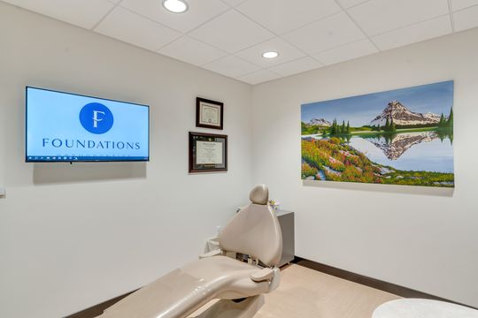 A Foundations Orthodontics exam room featuring a treatment chair, wall-mounted brand display, credentials, and colorful mountain artwork.