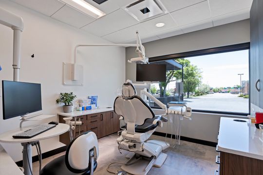 A clean and modern treatment room at Arvada Dental Center featuring a patient chair, dental equipment, wall-mounted monitor, computer station, wood cabinetry, and a large window with natural light.