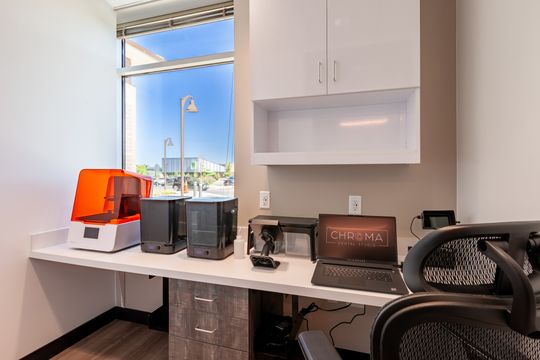 This image of the office workstation with city view at Chroma Dental Studio shows a desk, computer equipment, cabinetry, window light, chair, and compact office layout.