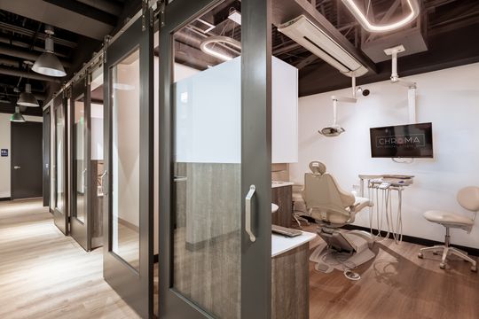 This image of the glass treatment suite hallway at Chroma Dental Studio shows glass partitions, frosted panels, dental chairs, overhead lights, and exposed ceiling design.