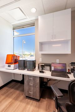 This image of the vertical office workstation view at Chroma Dental Studio shows a desk, chair, white cabinetry, window light, and organized desktop equipment.