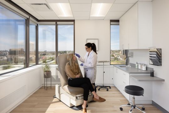 This image of the dermatology consultation room at Core Dermatology shows a clinician with a seated patient, exam chair, wall monitor, large windows, and city views.