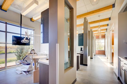 This image of the hallway and treatment room view at Meridian Periodontics of Central Park shows operatories, glass partitions, wood beams, wall monitor, and natural light.