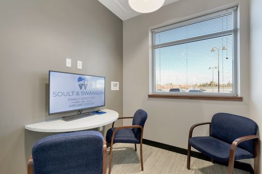 This image of the consultation room with monitor at Soult and Swanson Family Dentistry shows a wall monitor, round table, blue chairs, window view, and compact patient consultation setup.