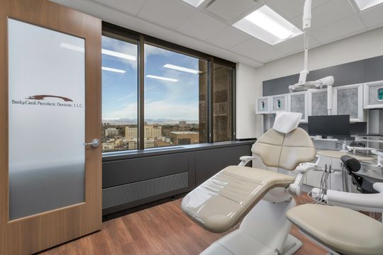 A bright treatment room at Bridge Creek Dentistry featuring a modern dental chair, large windows, mountain and city views, digital monitors, and clean contemporary clinical finishes.