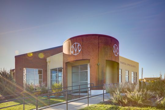 This image of the sunlit exterior view at Meridian Periodontics of Central Park shows front windows, curved wood entry, accessible ramp, exterior signage, and warm sunlight.
