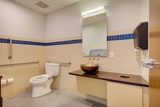 This image of the accessible restroom at Meridian Periodontics of Central Park shows toilet, sink, mirror, blue tile accent, grab bars, and clean restroom finishes.