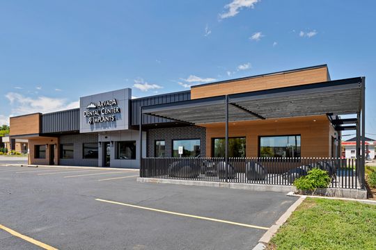 An exterior view of Arvada Dental Center & Implants in Colorado, showing the modern building facade, prominent signage, wood-look exterior panels, black accents, parking area, and covered patio.